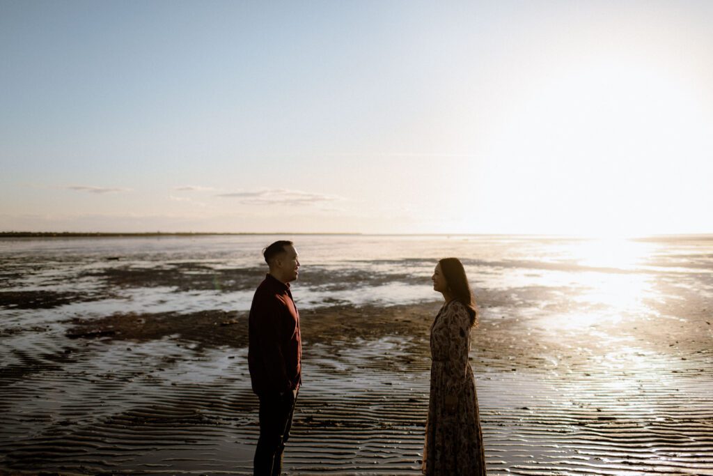 A couple smiling at each other in front of a sunset at a beach during their engagement session in Vancouver.