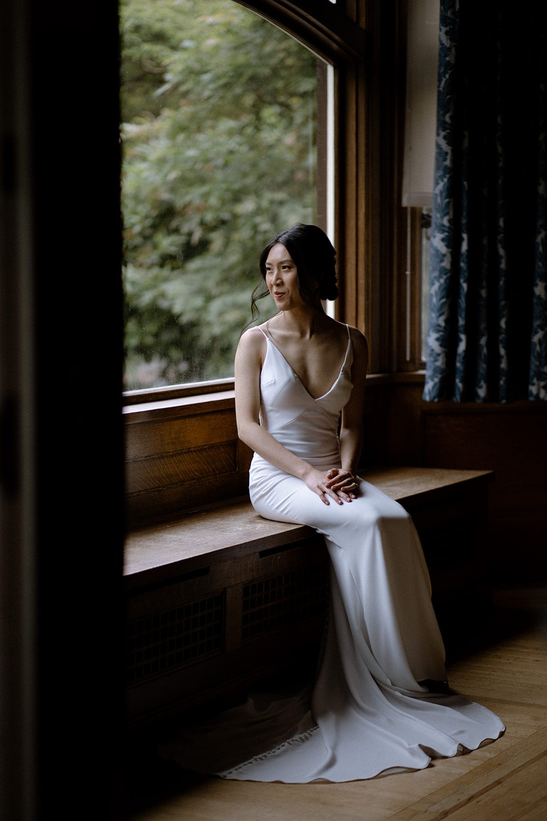 Bride sitting on a bench by a large window looking out of the room at Cecil Green Park House.