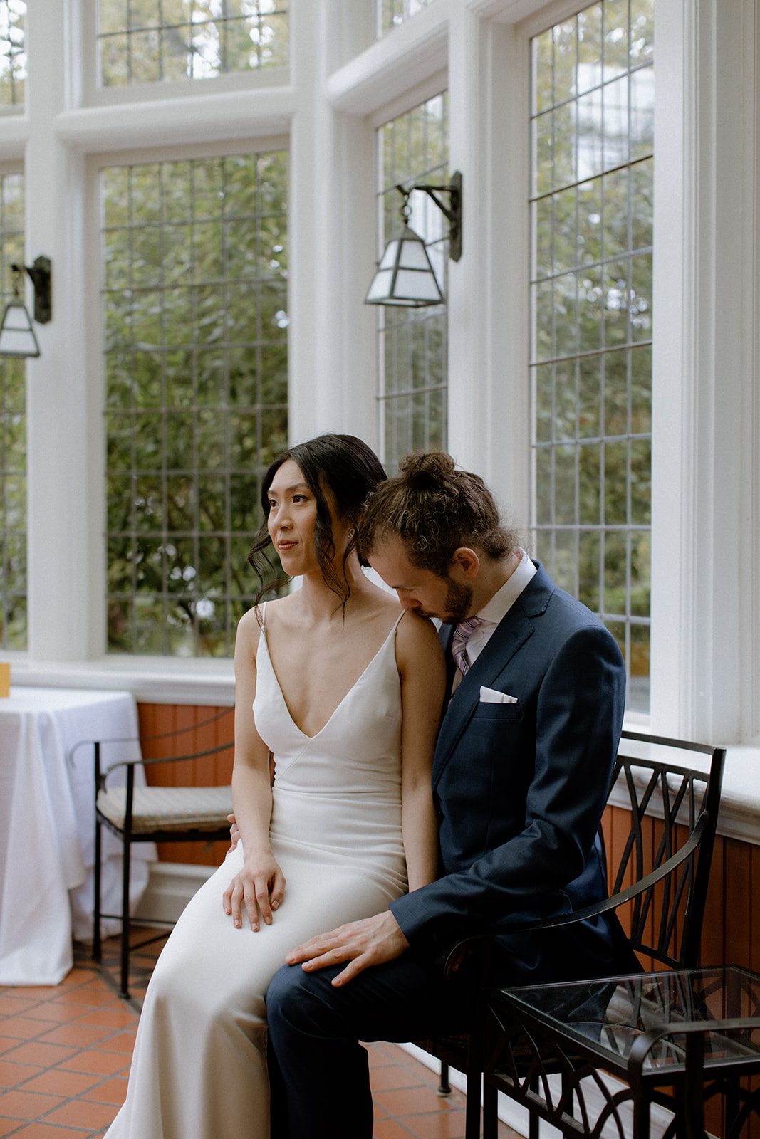 Bride sitting on groom's lap while he kisses her shoulder in a sunroom at Cecil Green Park House.